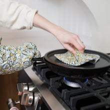 Load image into Gallery viewer, Image shows the oven mitt and potholder in use. Model is holding a cast iron pan with oven mitt while taking the lid off the pan using the pot holder. 